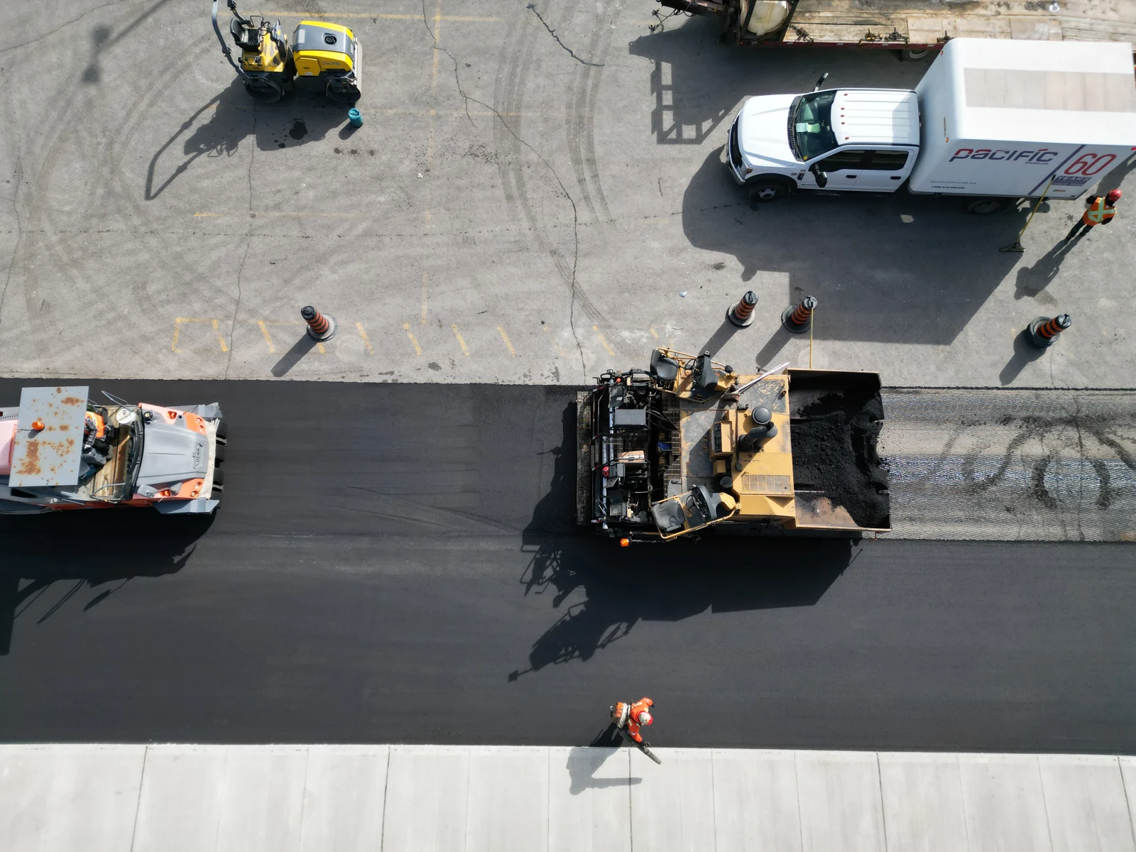 Drone view of a paving crew, asphalt paver, roller, and Pacific Paving truck beside a newly paved strip.