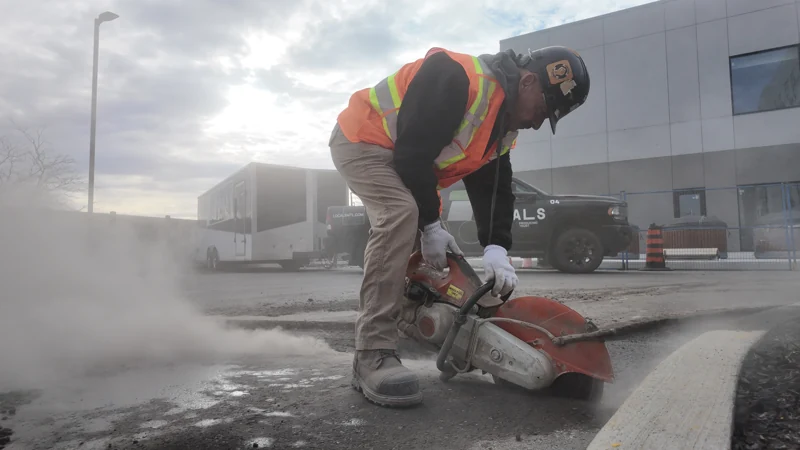 A crew member uses a handheld pavement saw to cut asphalt at a commercial worksite.