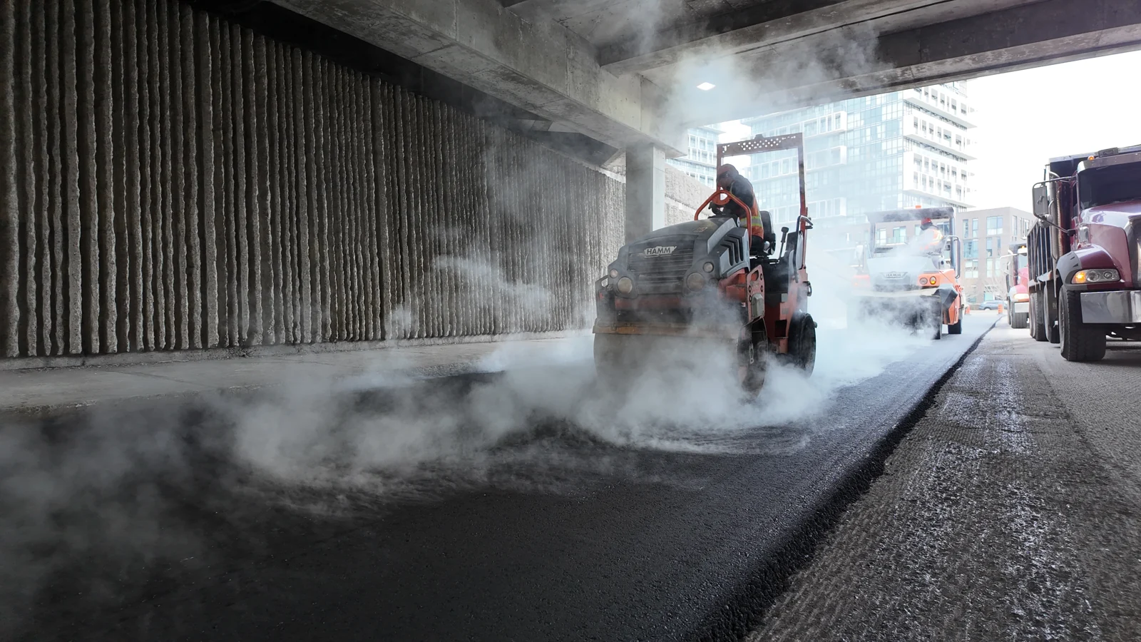 A roller compacts fresh asphalt under a bridge, with steam rising from the new pavement.